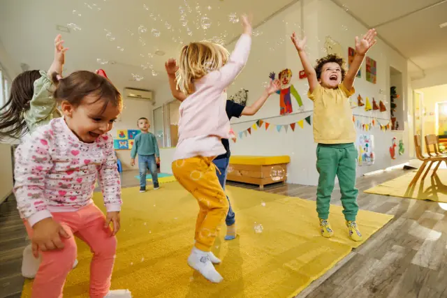 Toddlers jumping with their arms in the air as bubbles float around a bright playroom.