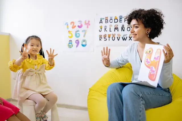A teacher holding up a number card while a young girl in a yellow dress counts on her fingers, with number and alphabet posters on the wall behind them.