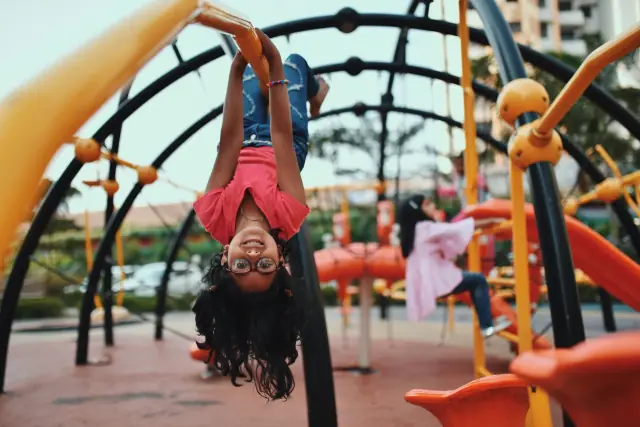 A girl hanging upside down and grinning on playground climbing bars, with other children playing behind her.